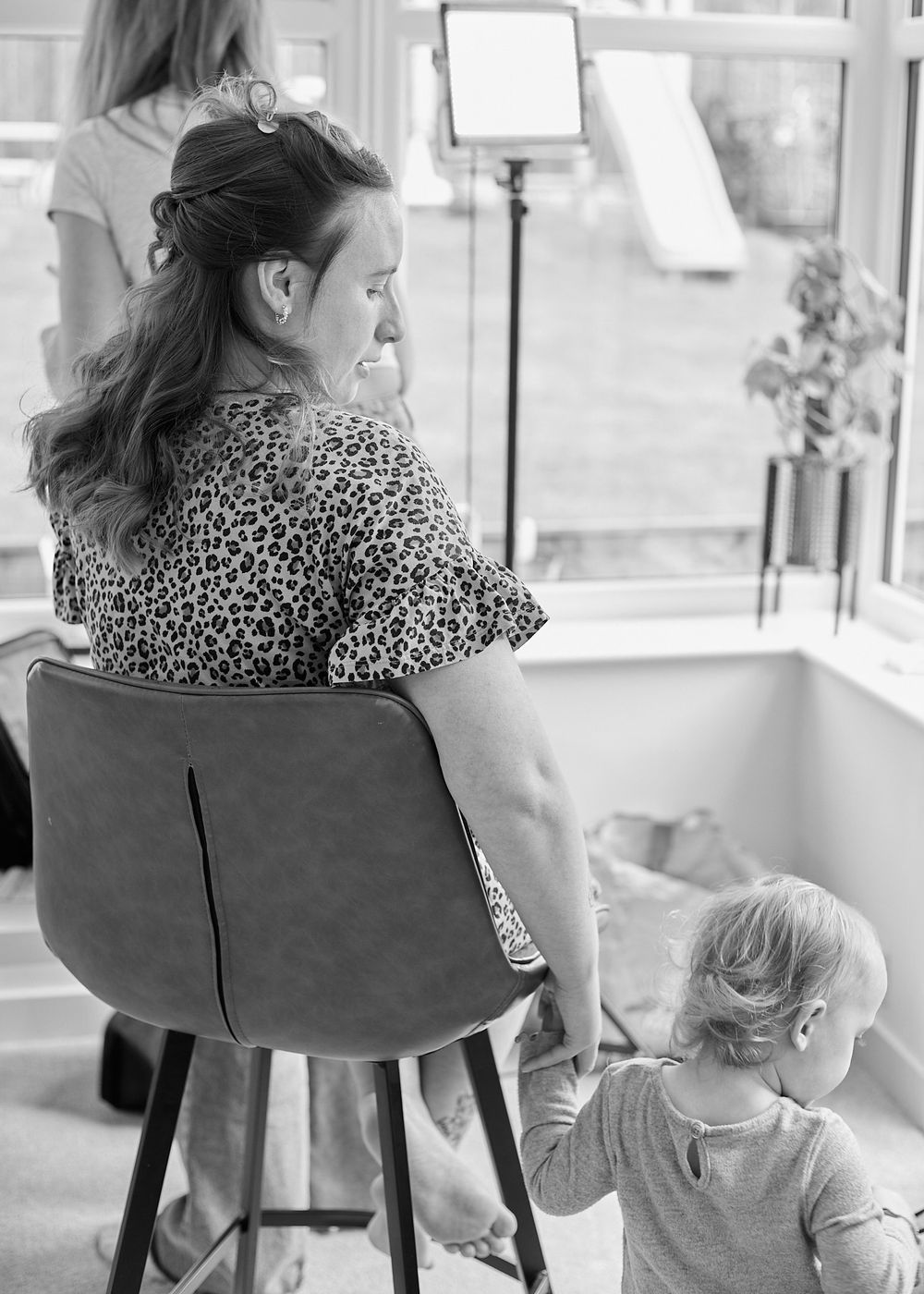 Black and white photo of a woman in leopard print seated on a stool while a toddler stands beside her.