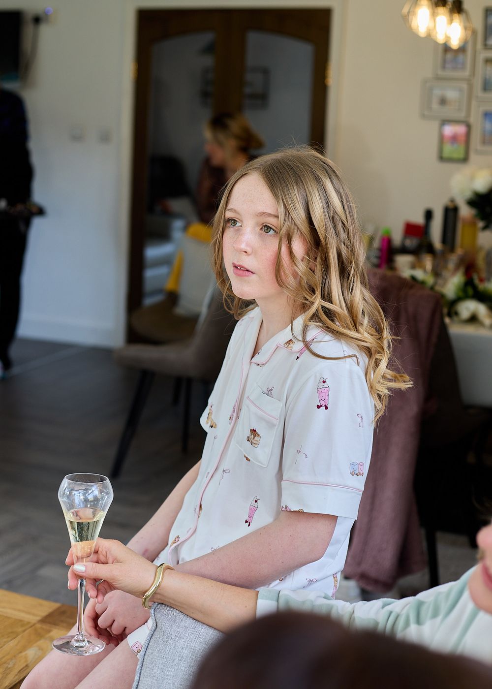 A young girl in a white patterned shirt holds a champagne flute while seated at a decorated table.