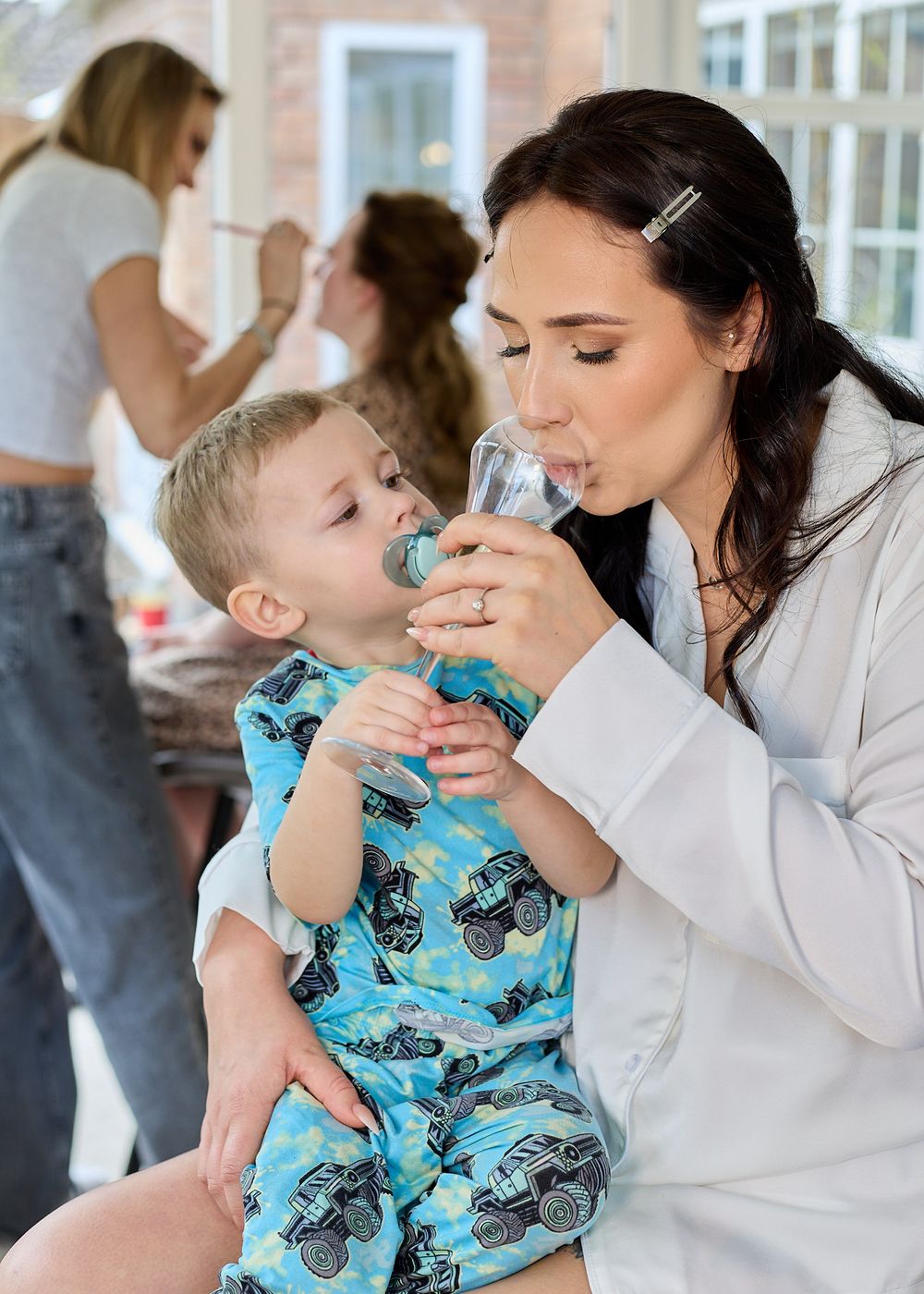 A dark-haired woman in a white robe shares a champagne glass with a toddler in blue monster truck pajamas.