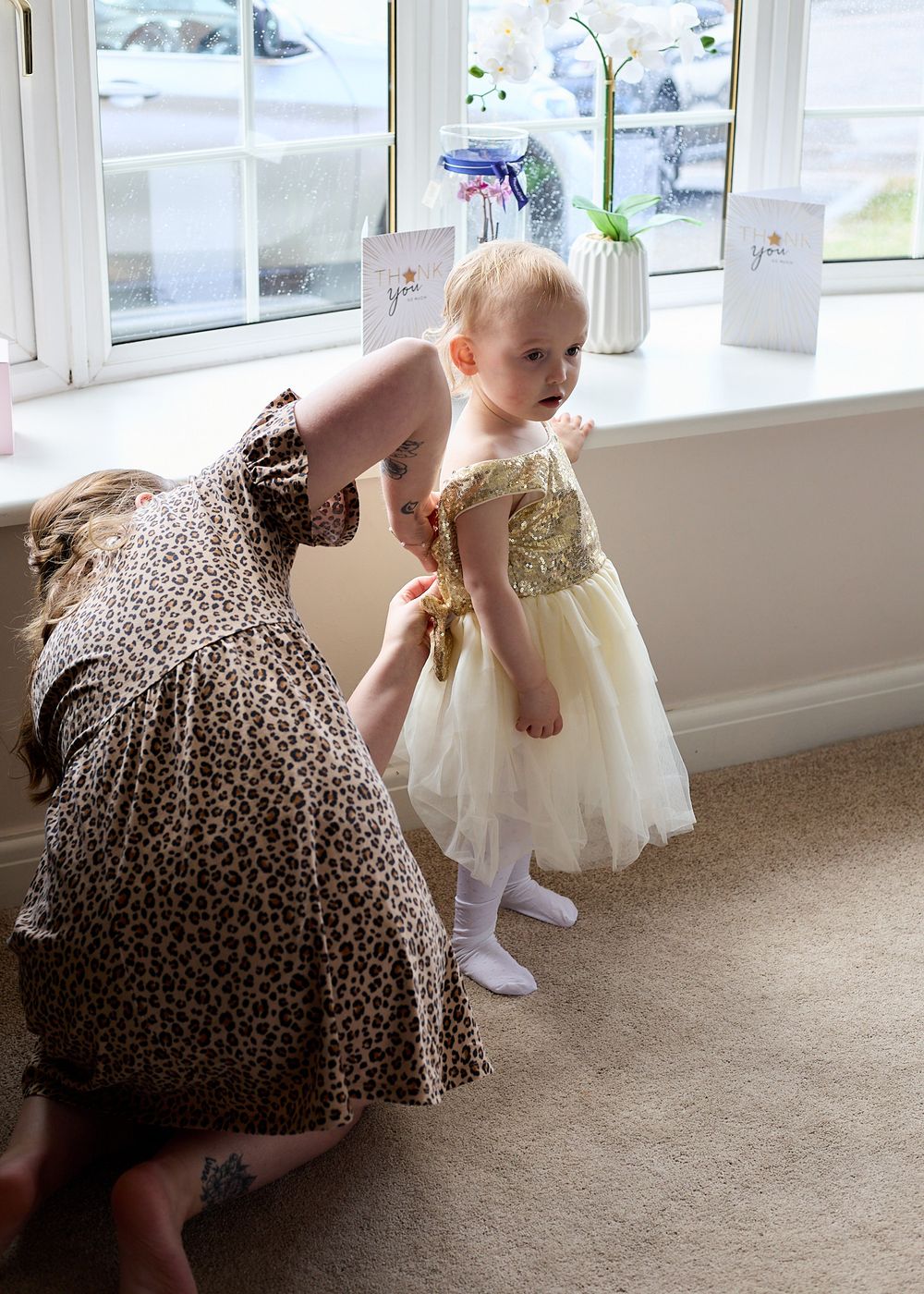 A woman in leopard print dress adjusts the golden tulle dress of a small toddler standing by a window.