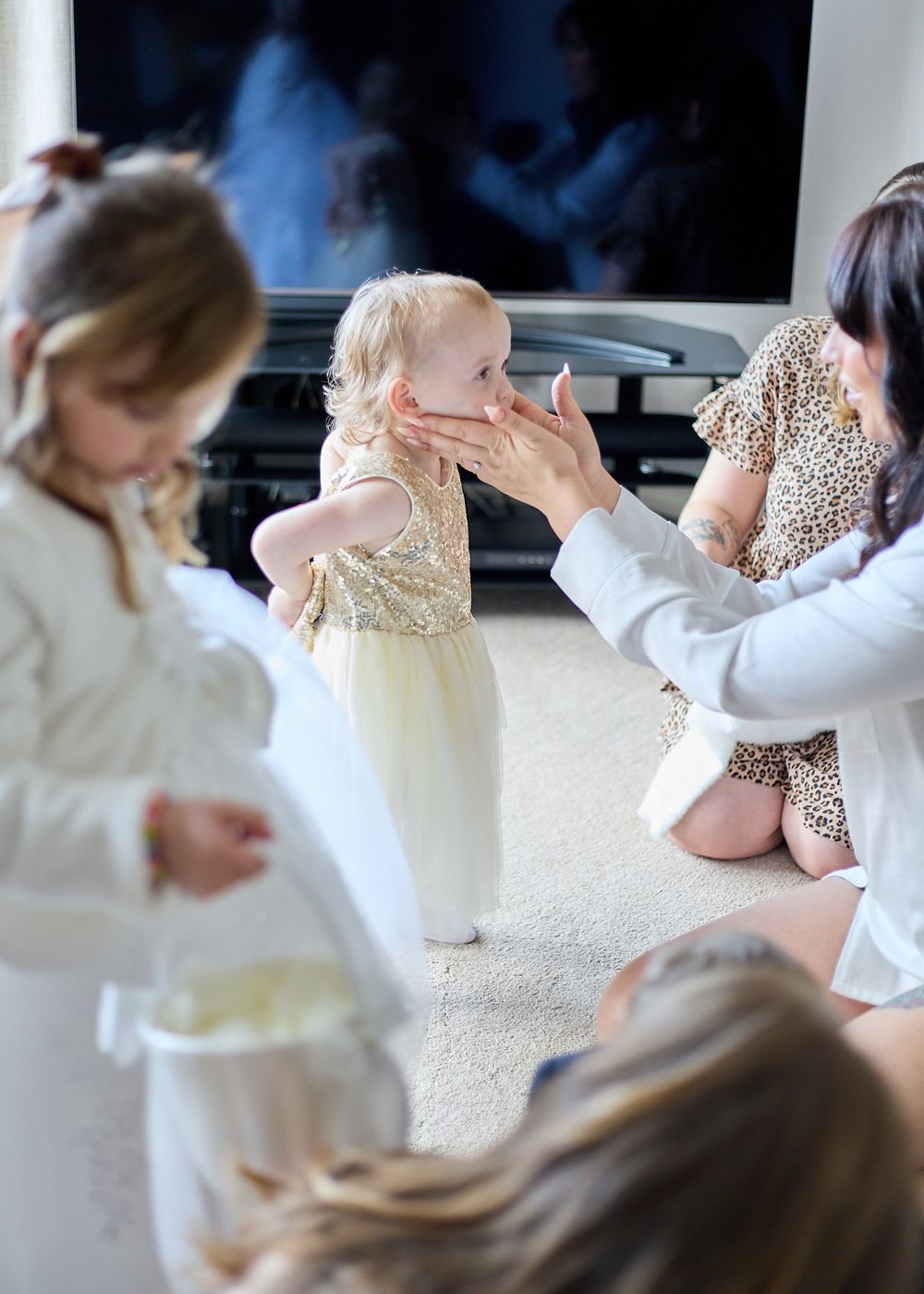 A woman kneels to touch a baby girl in a gold dress during wedding preparations indoors.