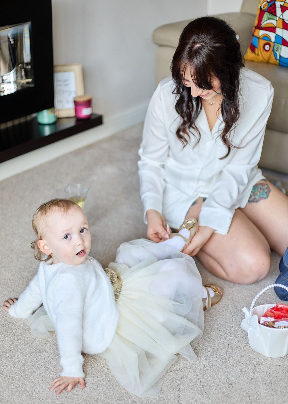 A dark-haired woman in a white robe helps dress a baby in a tulle skirt on the carpet.