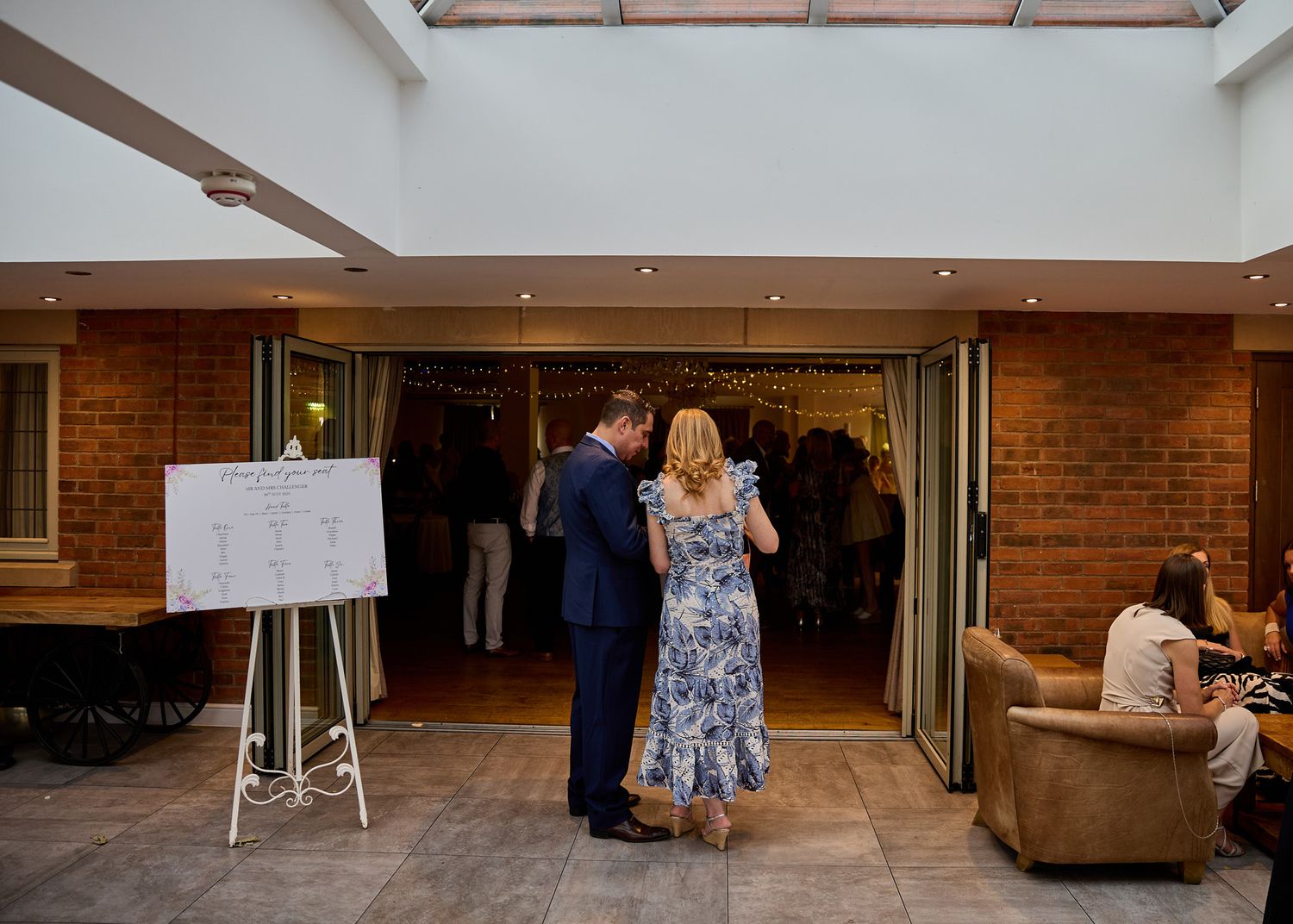 Couple reviewing seating chart on easel at elegant wedding venue with brick walls and skylight.