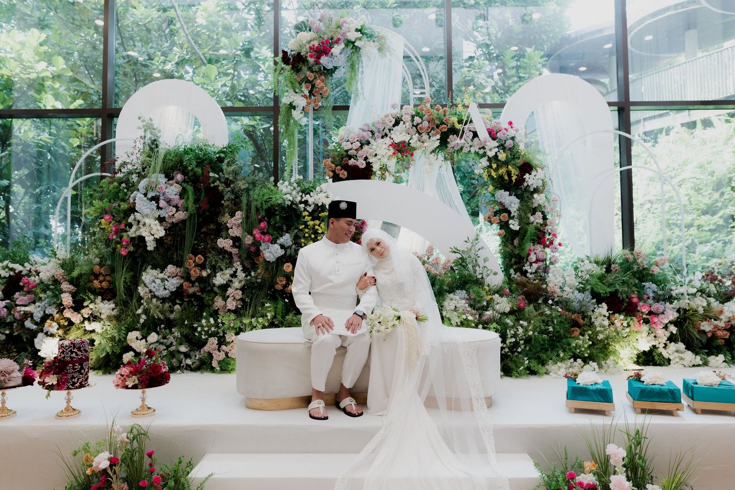 Muslim couple in white wedding attire seated at floral decorated altar with lush greenery and elegant flower arrangements.