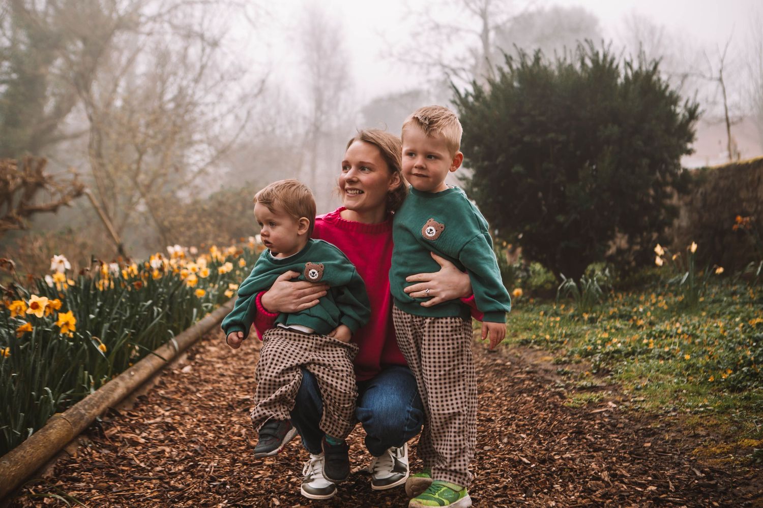 A mother crouches with two young boys in matching outfits among daffodils in a misty garden.