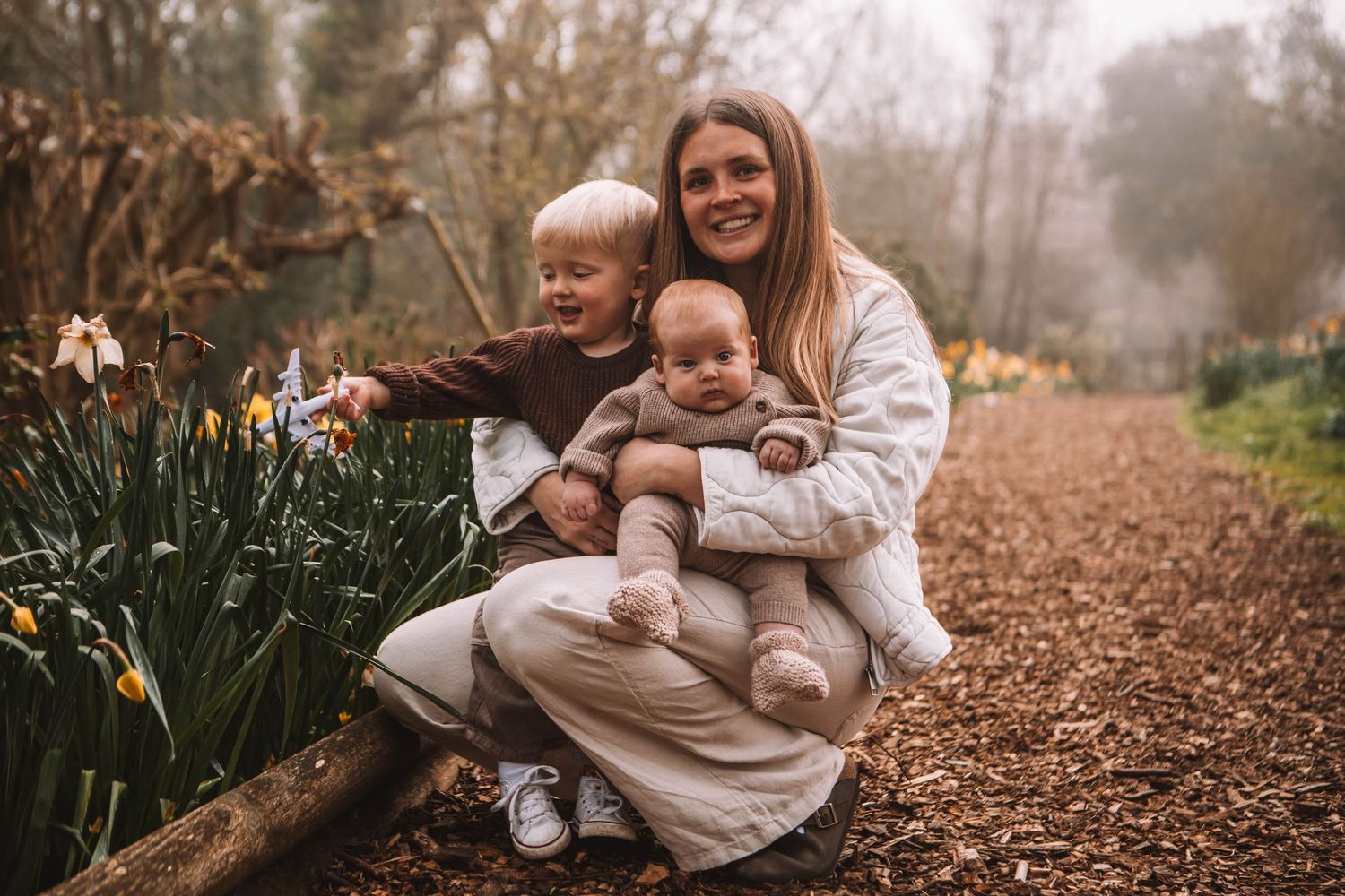A smiling woman crouches with two young children beside spring daffodils on a garden path.