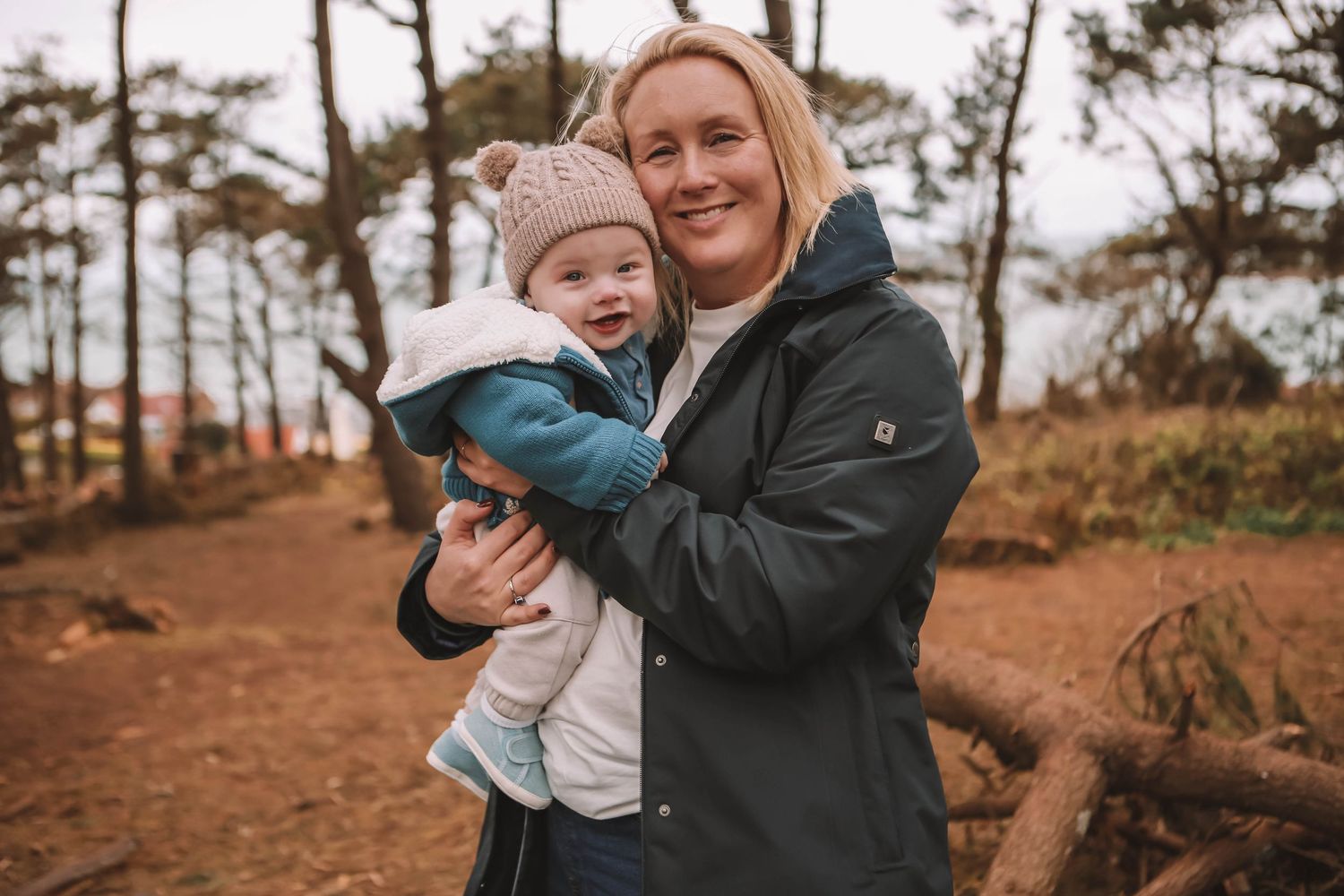 A smiling woman in a dark jacket holds a happy baby in a knit hat outdoors in a forest setting.