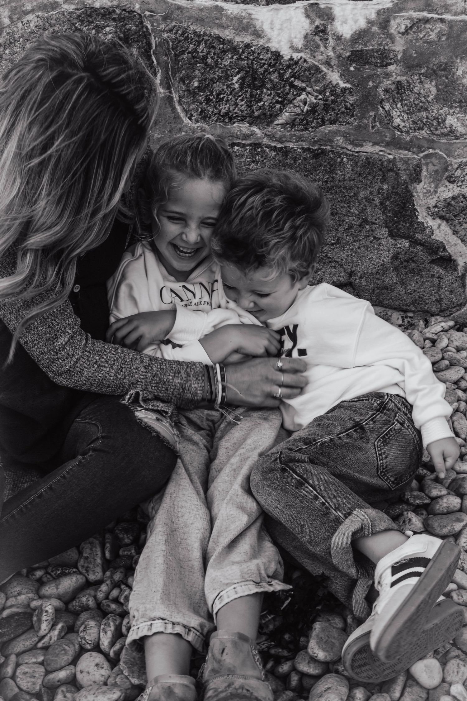 Black and white photo of a mother hugging two young children sitting on pebbles, all laughing joyfully outdoors.