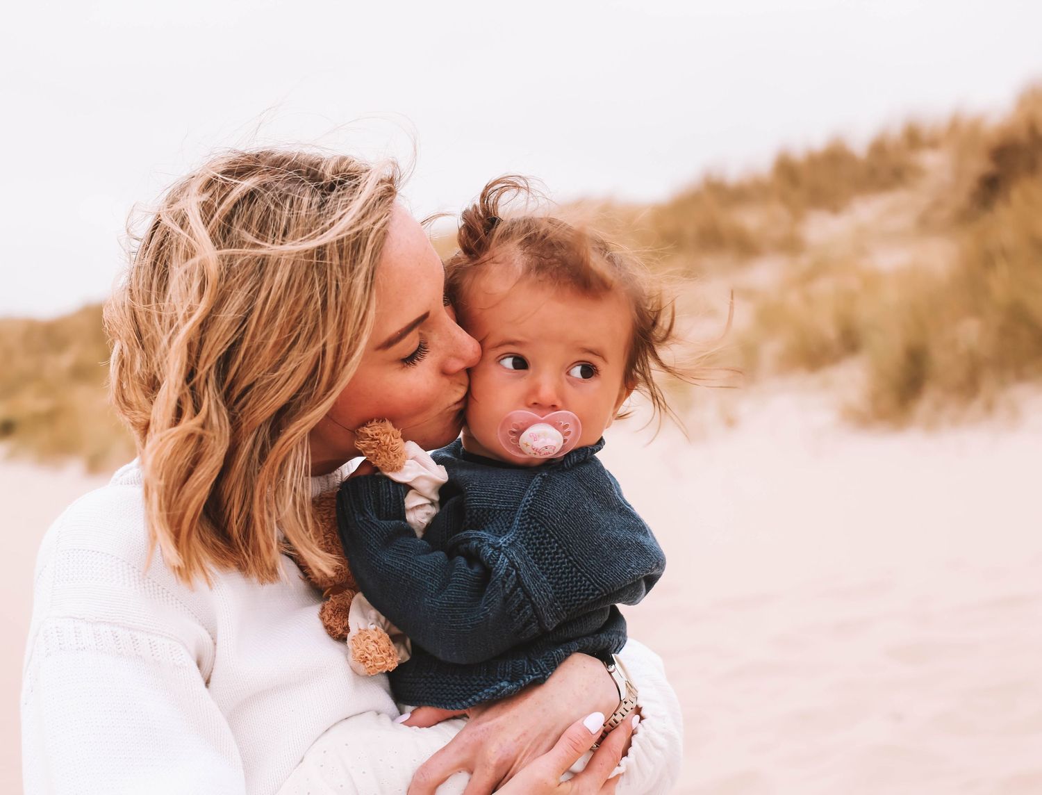 A loving mother kisses her baby on the cheek at a sandy beach, baby wearing denim jacket with pacifier.