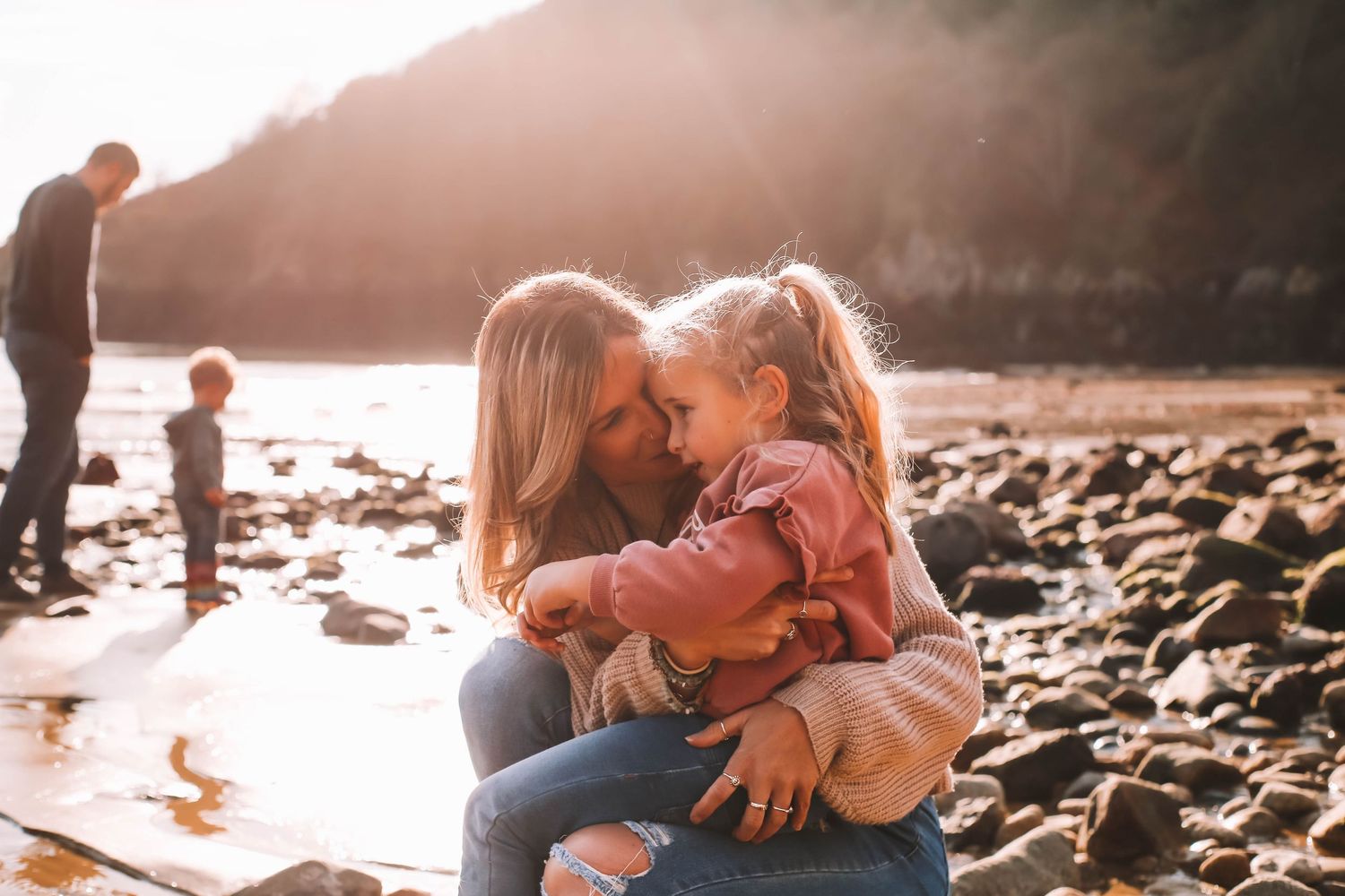 A mother and young daughter share a tender forehead-to-forehead moment on a rocky beach at golden hour.