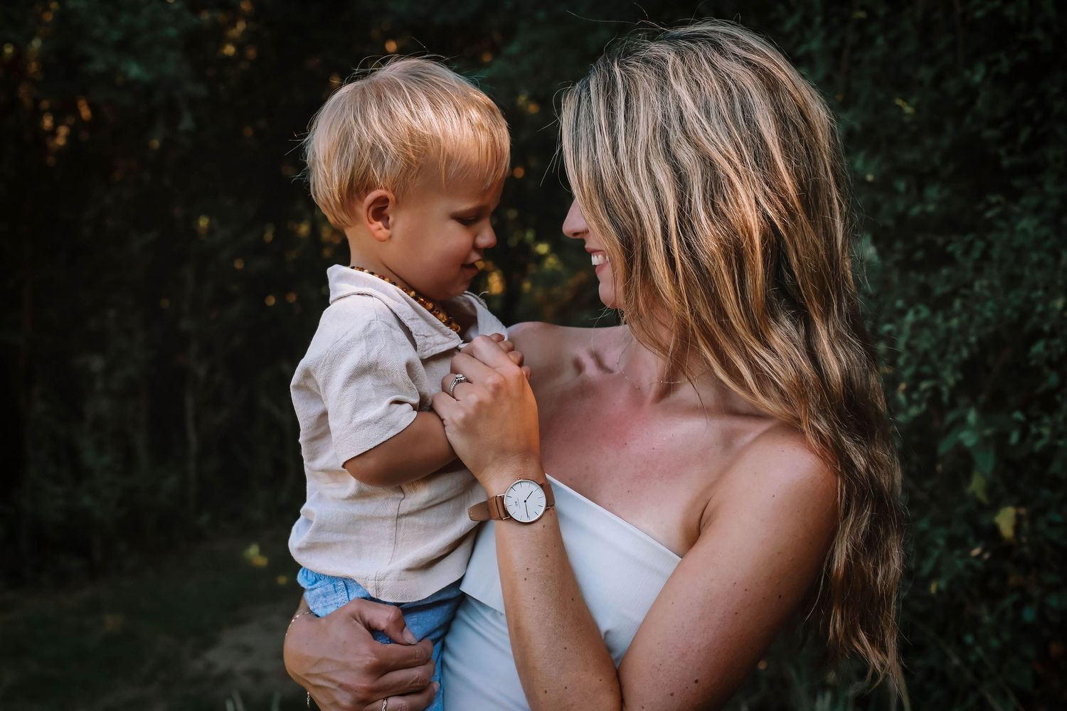 A smiling mother with long blonde hair holds her toddler son outdoors, sharing a tender moment together.