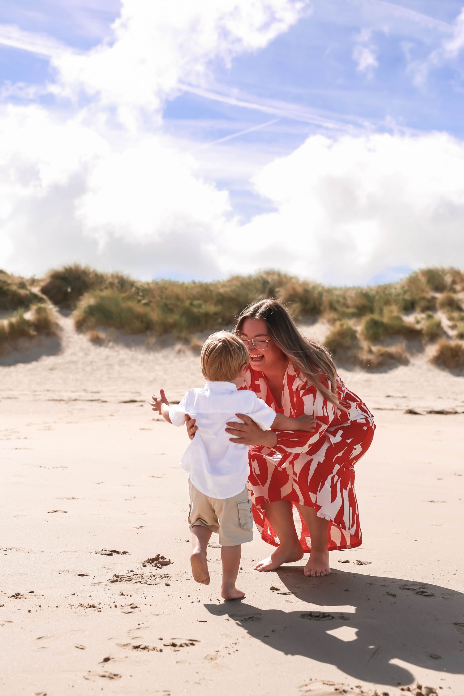 A laughing mother in a red floral dress crouches to embrace her toddler son on a sandy beach with dunes behind them.