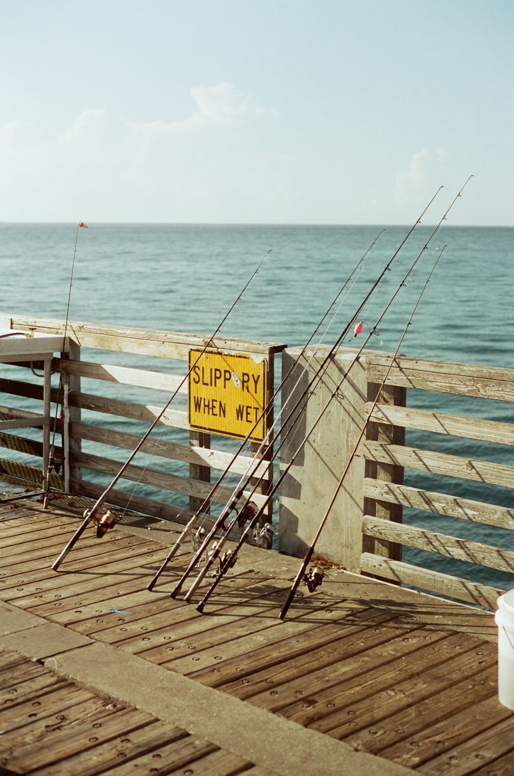Fishing rods leaning against a wooden pier railing with a yellow slippery when wet sign and ocean view.