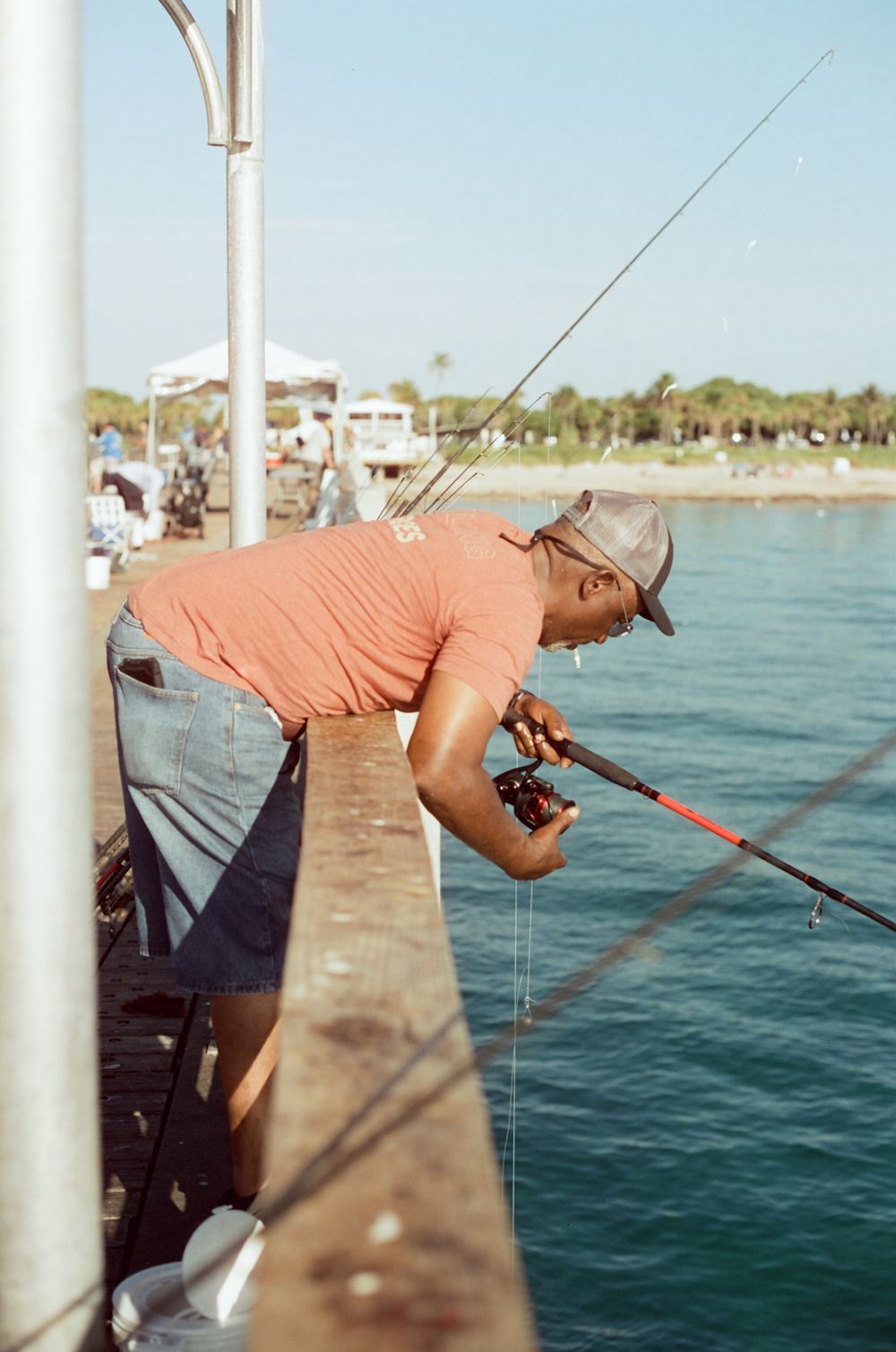 Man in orange shirt and cap fishing from a pier over calm blue water on a sunny day.