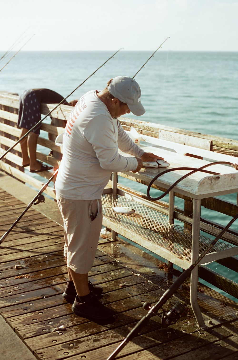 Man in gray cap and white shirt fishing on a wooden pier with rods and ocean in background.