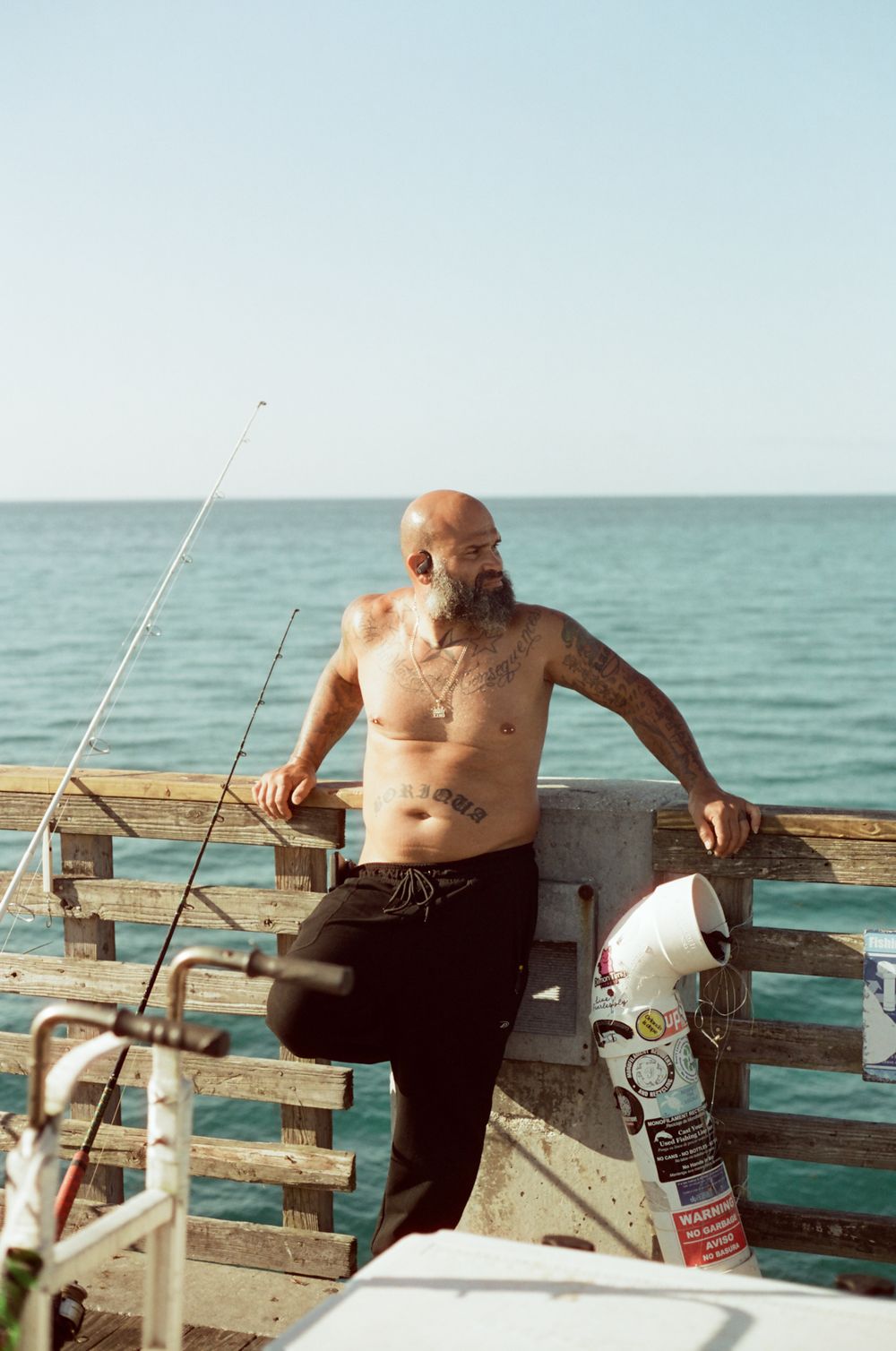 Tattooed shirtless bald man with beard leaning on pier railing beside fishing rods and ocean.
