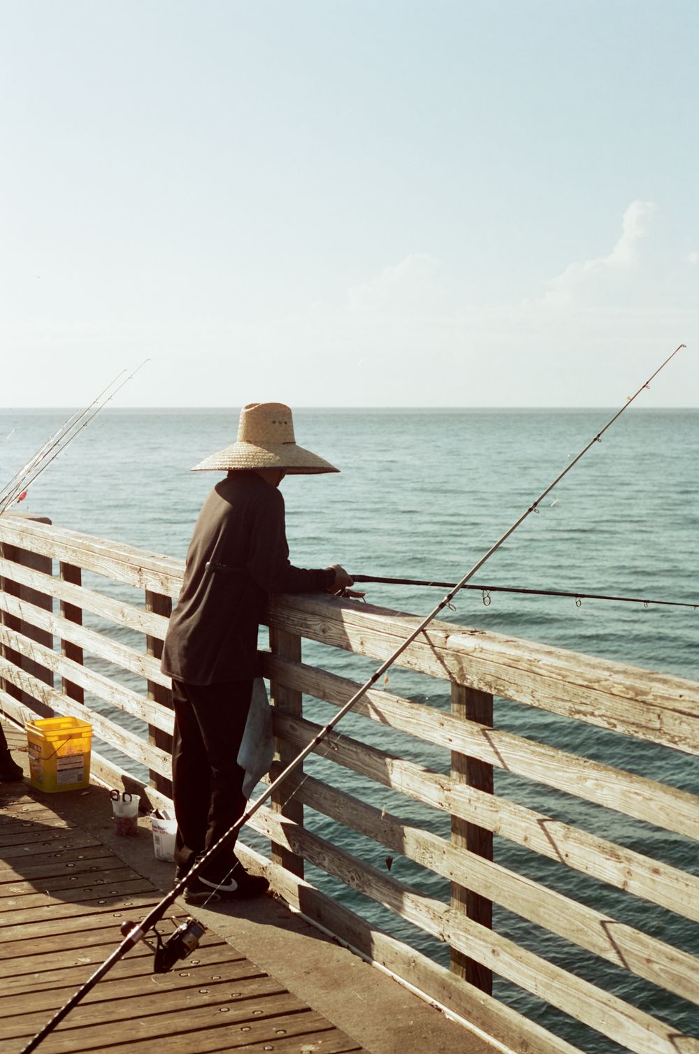 Lone fisherman wearing wide-brim hat casting two rods from a wooden pier overlooking the ocean.