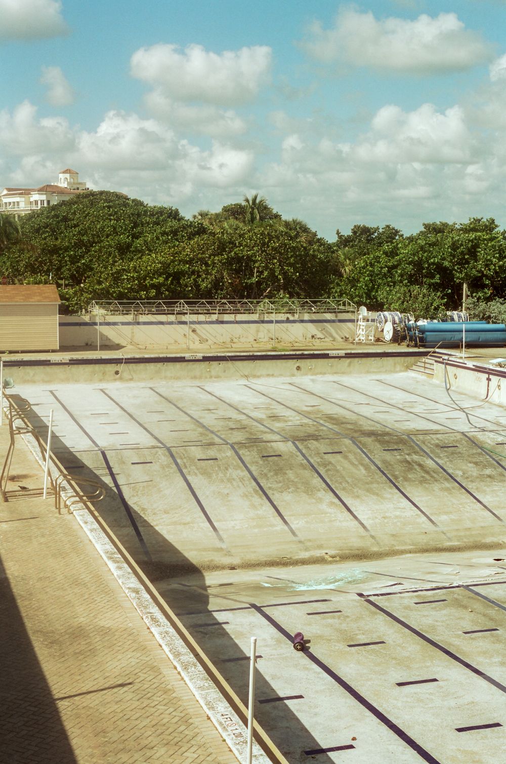 Empty outdoor swimming pool with lane markers under a partly cloudy sky surrounded by trees.