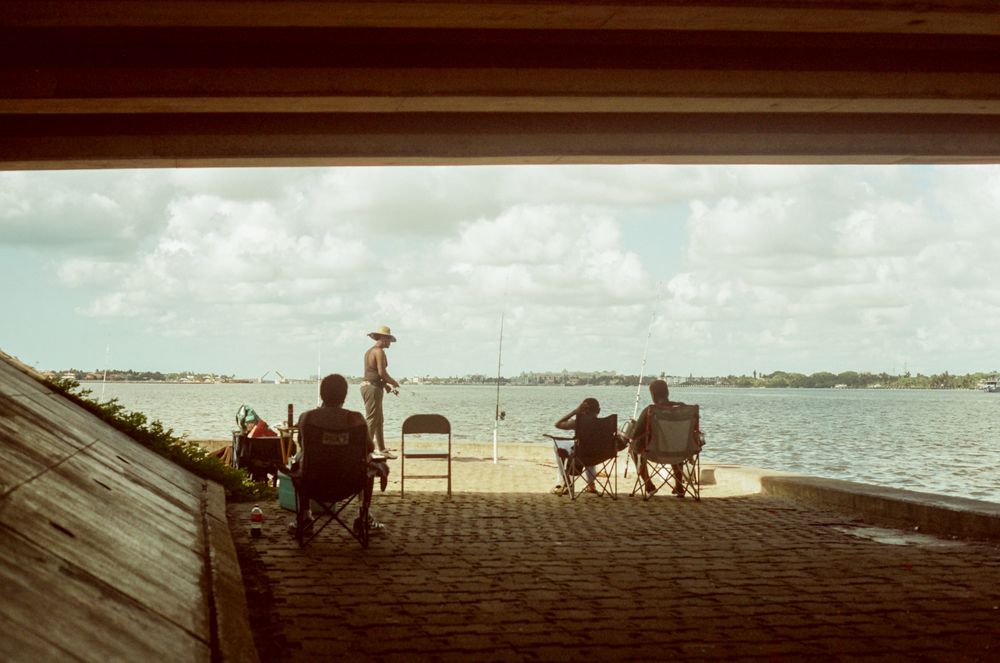People relaxing in chairs under a covered dock, overlooking a calm bay with sailboats on a cloudy day.