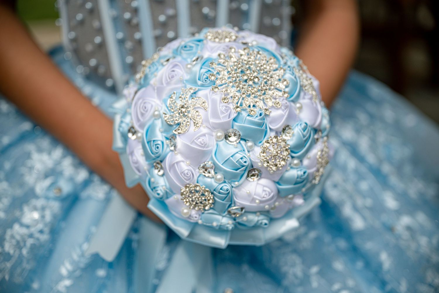Light blue bridal bouquet with satin ribbon roses and pearl brooch accents held by bride in matching lace gown.