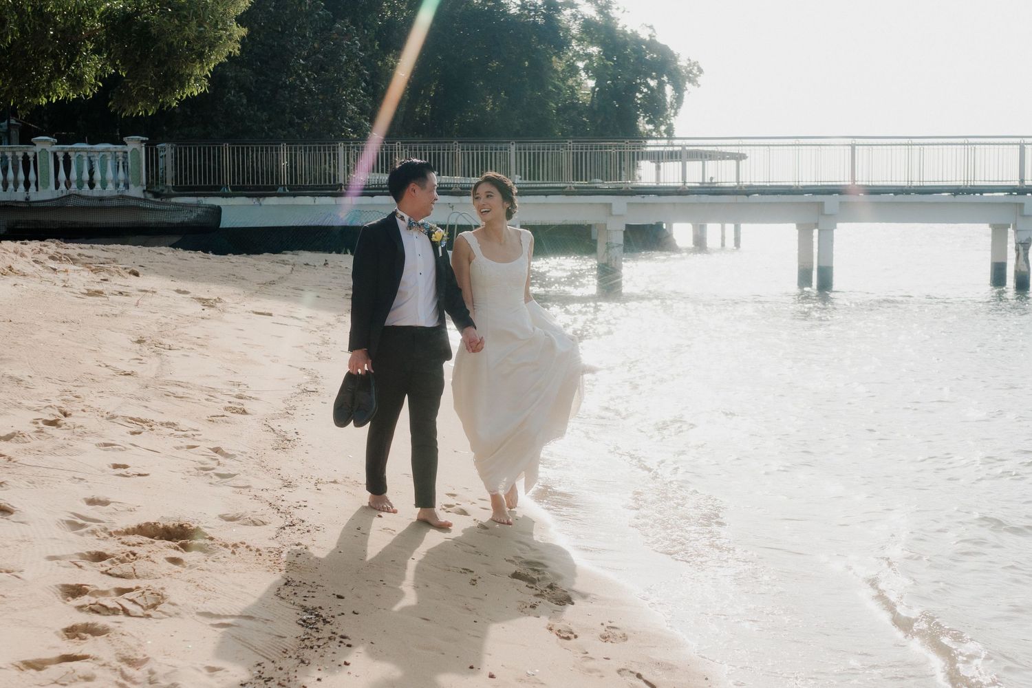 Bride and groom walking hand in hand on a sandy beach near a pier, wearing wedding attire in soft sunlight.