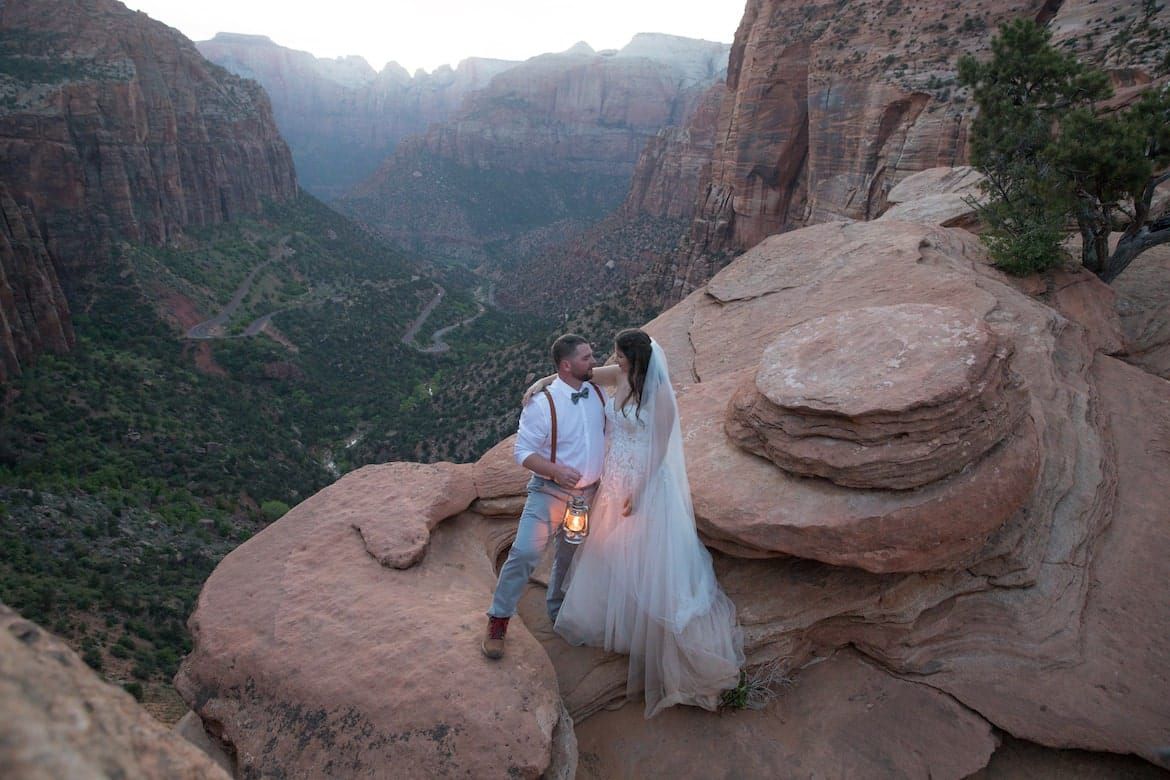 Bride and groom sit on red rock cliff edge at Zion National Park canyon overlook during sunset elopement ceremony.