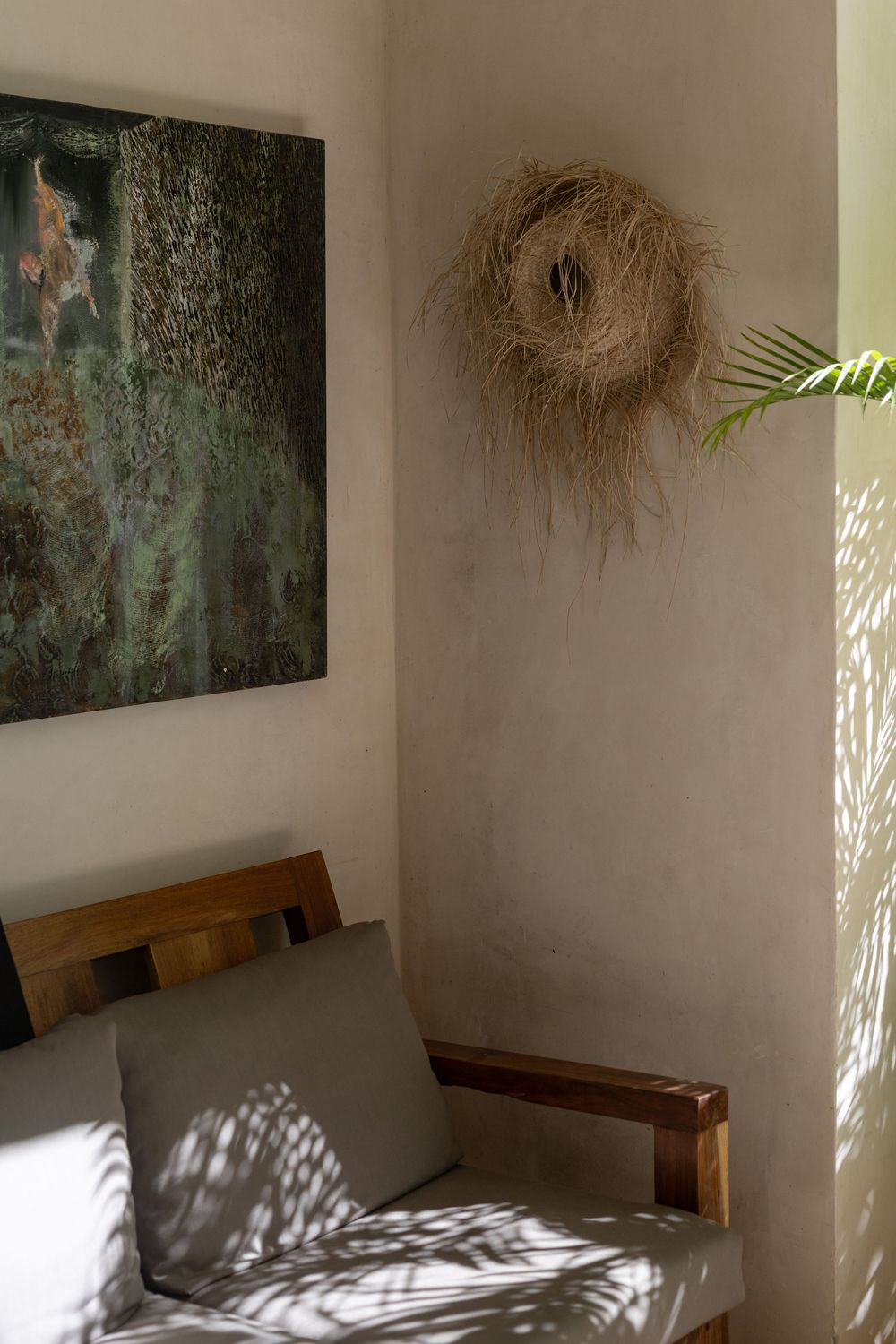 Minimalist room corner with wooden chair, green cushion, nature painting, and woven straw wall decor with palm leaf shadows.