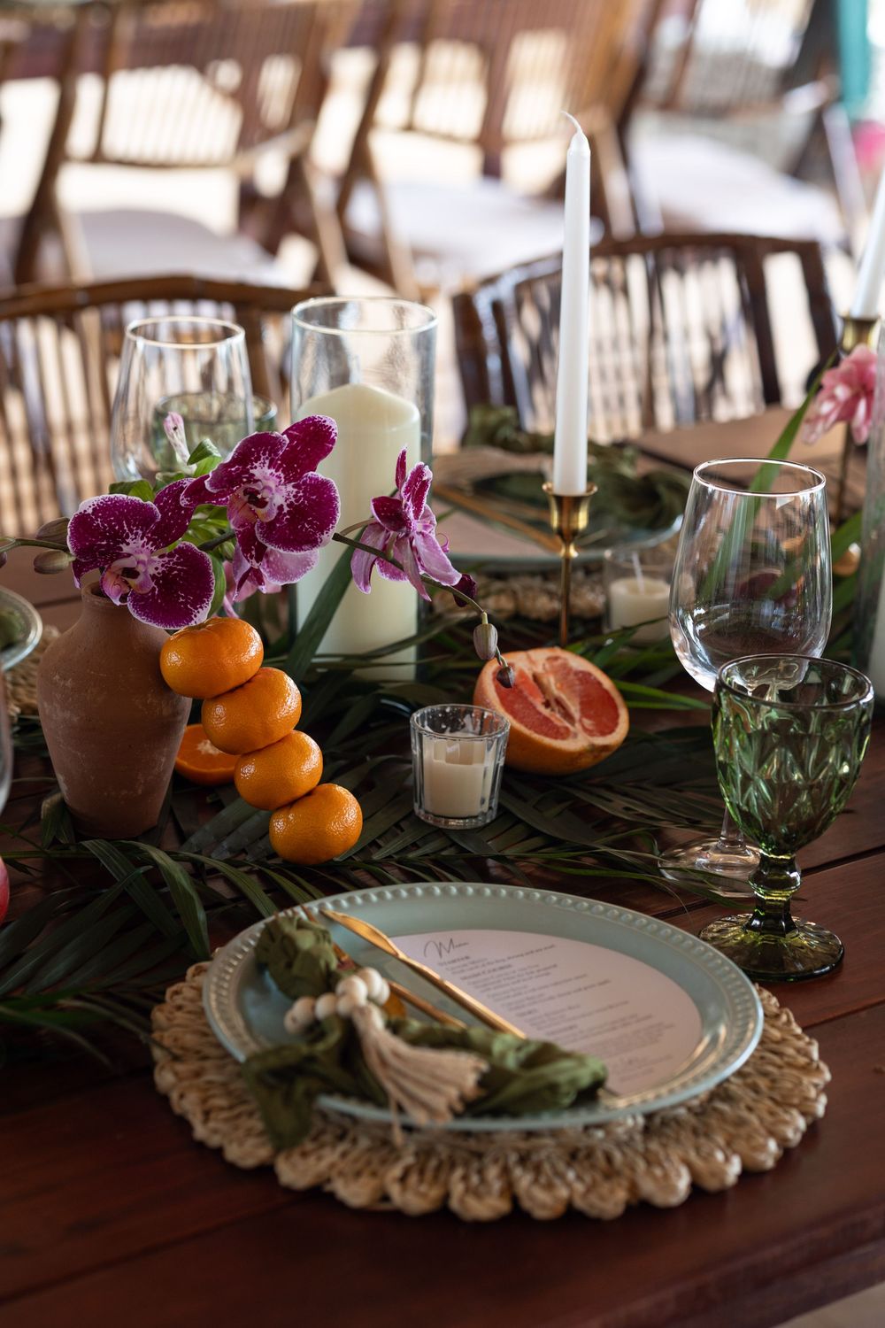 Elegant tropical table setting with orchids, oranges, grapefruit, candles, and decorative plates on woven placemats.