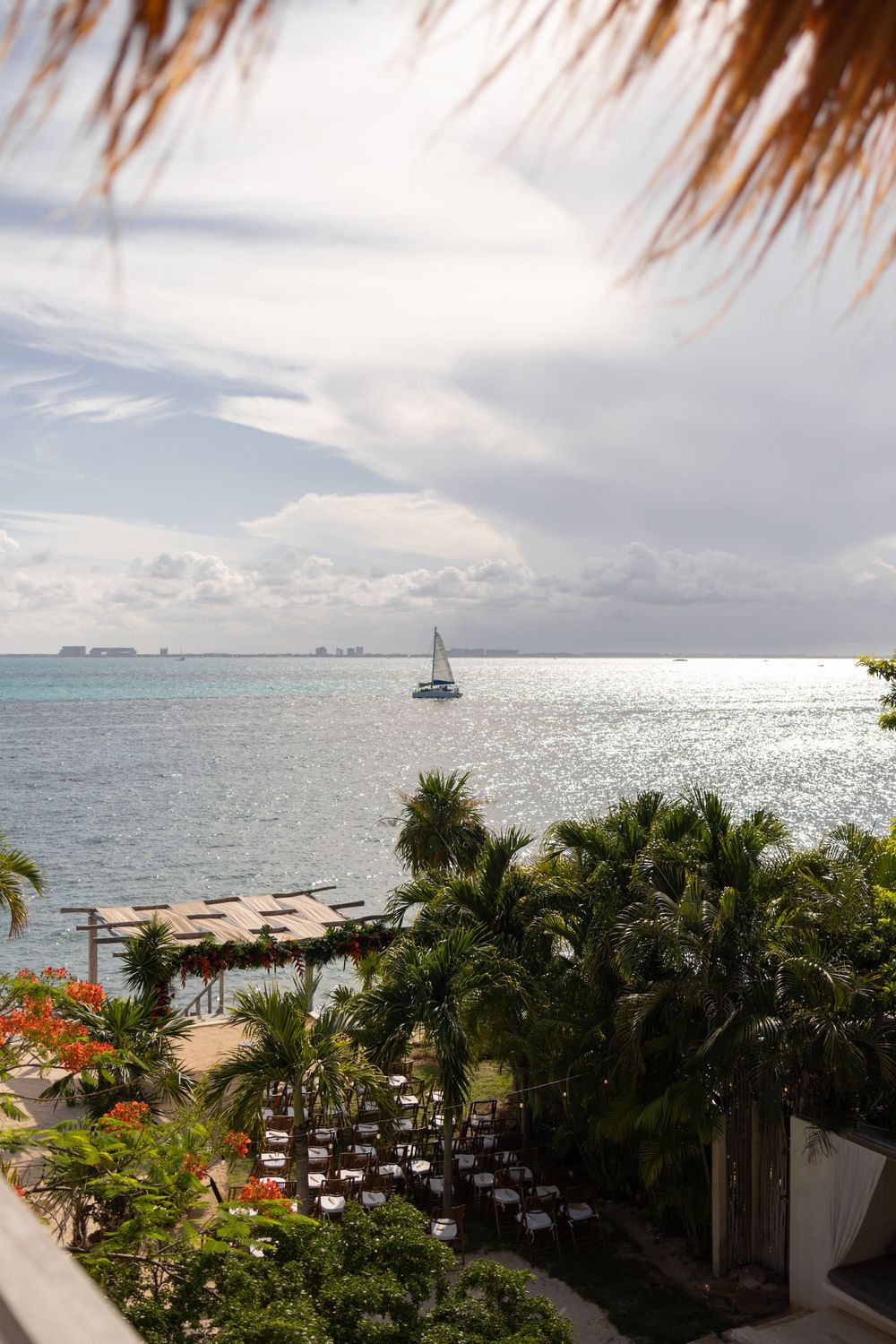 Tropical resort view with sailboat on calm Caribbean sea, palm trees, beach pier, and outdoor dining area under cloudy sky at Casa Coco.