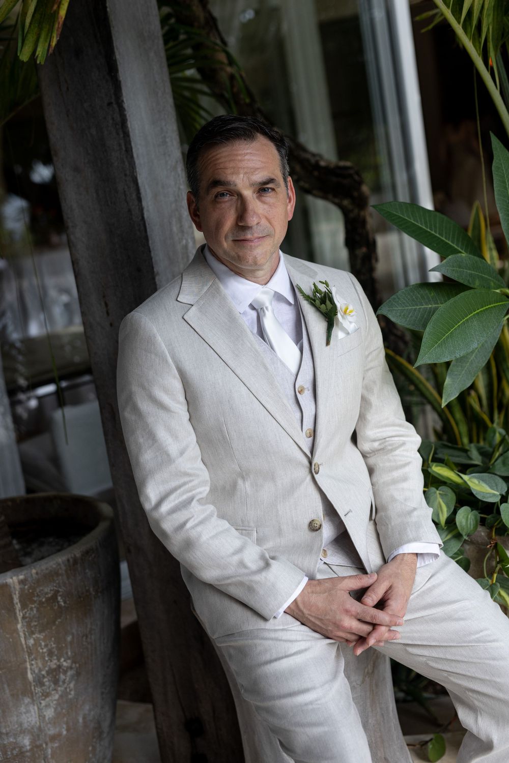 Elegant groom in cream linen three-piece suit with boutonniere, leaning against wooden post surrounded by tropical greenery.