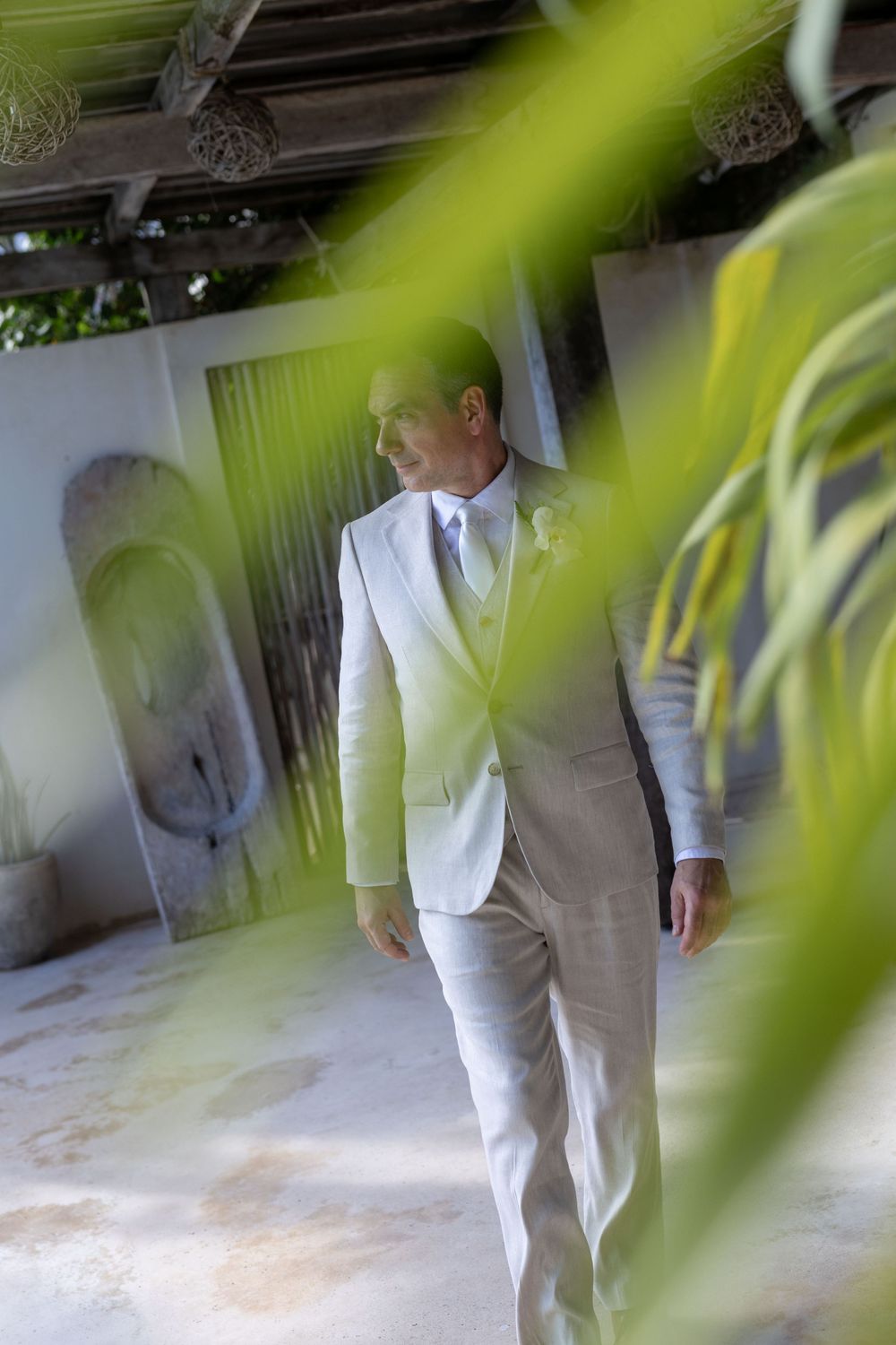 Elegant groom in white suit with boutonniere standing in tropical venue, photographed through lush green palm leaves.