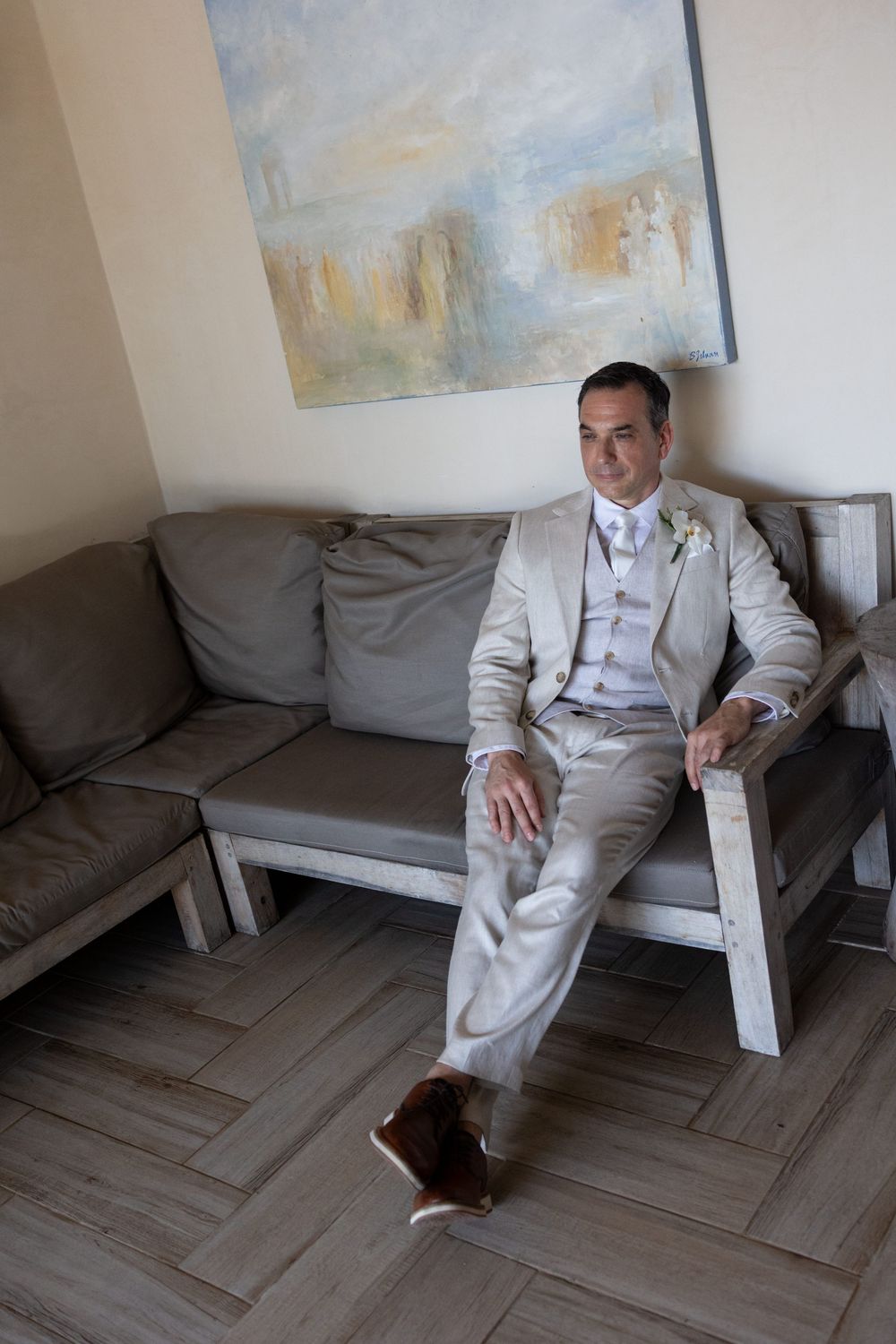Groom in beige suit with boutonniere sits relaxed on gray sofa, brown shoes, abstract painting behind him.