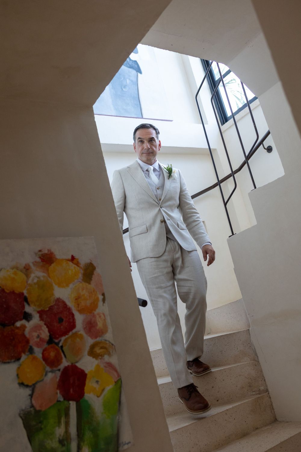 Groom in a light beige suit with boutonniere stands on stairs near floral artwork in elegant venue.
