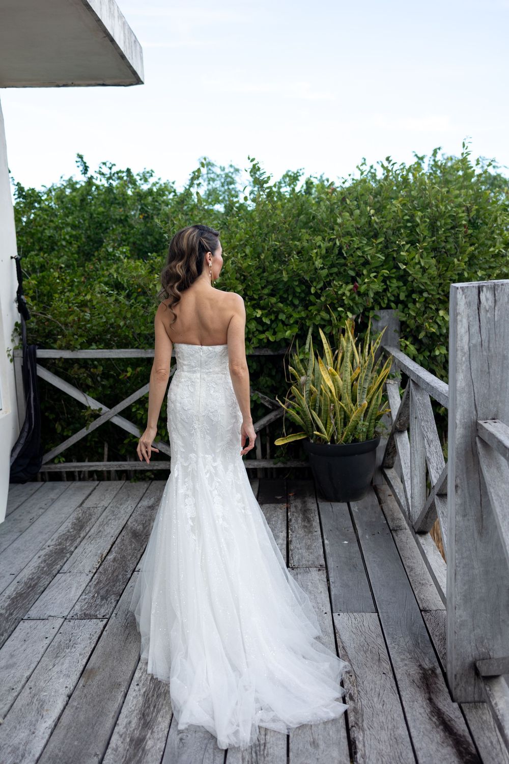 Bride in strapless lace mermaid wedding gown viewed from behind on wooden deck with green hedges.