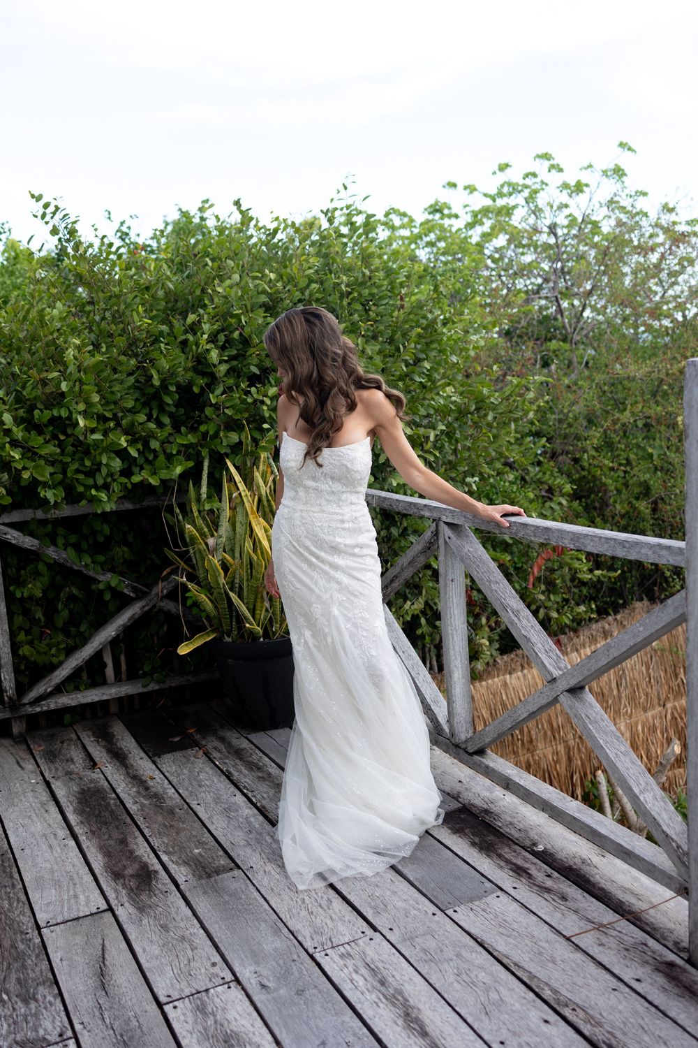 Bride in white lace wedding gown stands on rustic wooden deck overlooking lush green garden.