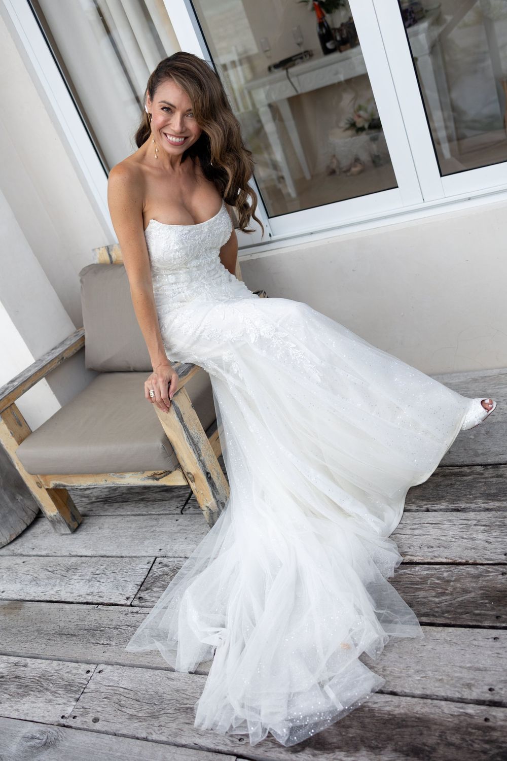 Smiling bride in a strapless white lace mermaid wedding gown sitting on a chair on a rustic wooden deck.