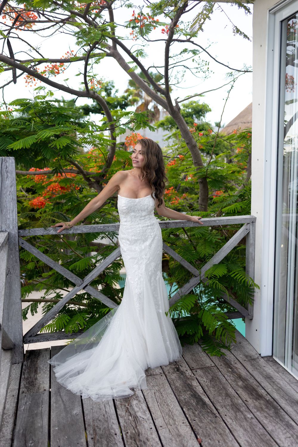 Bride in strapless white mermaid wedding gown standing on wooden deck surrounded by lush tropical greenery.
