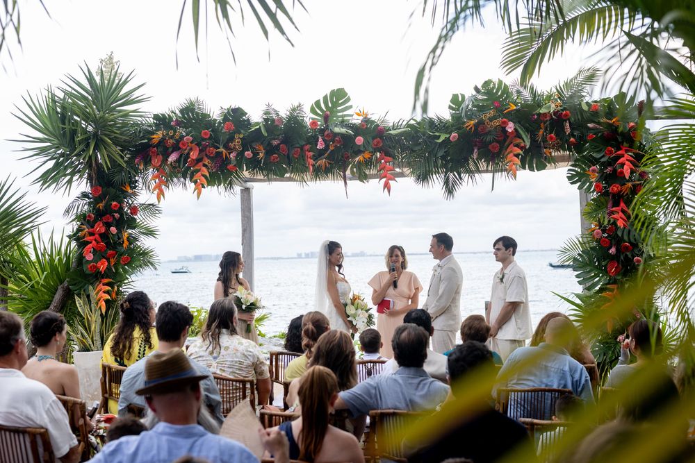 Tropical beach wedding ceremony under floral arch with ocean view, guests seated outdoors amid lush palm trees.