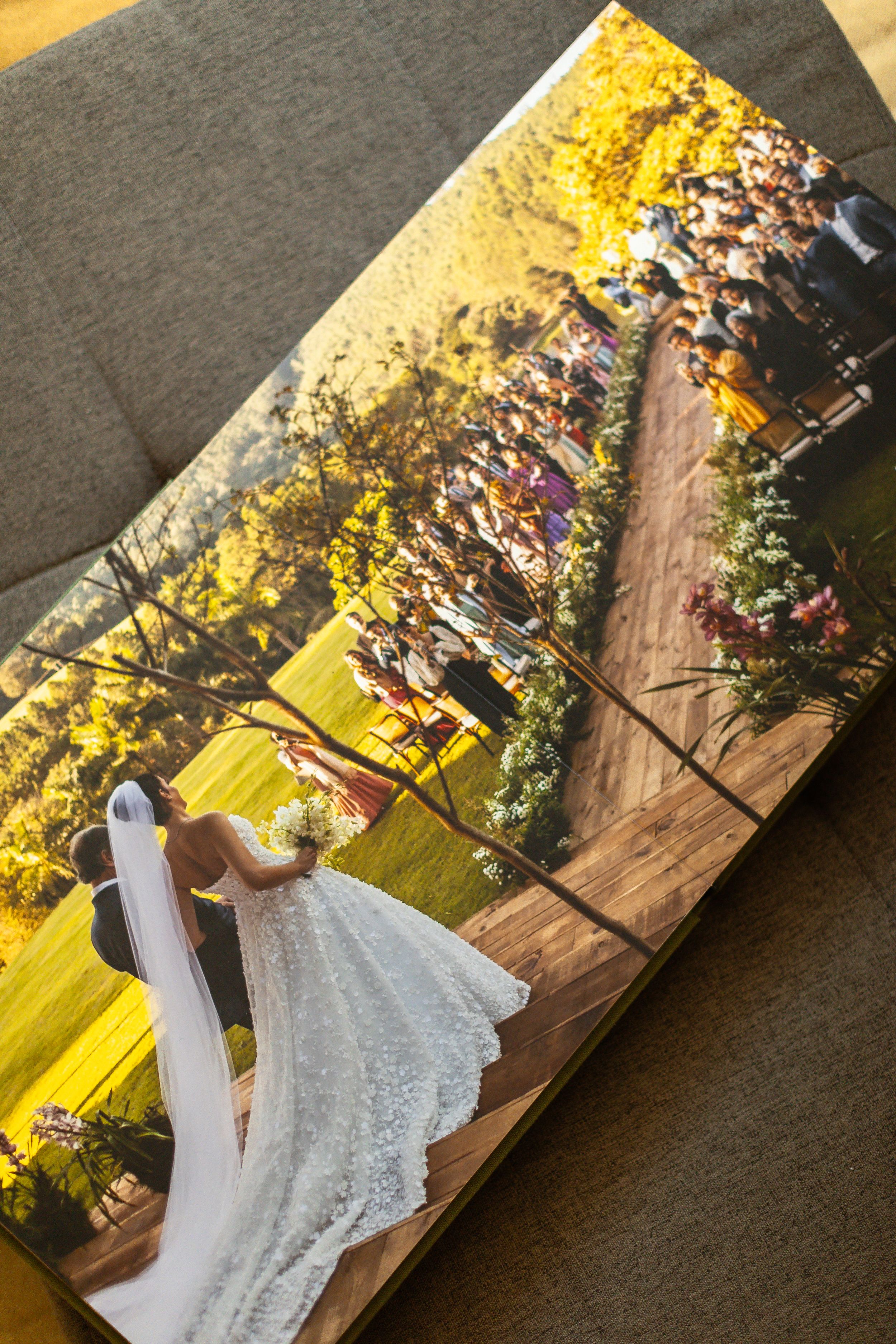 Open wedding photo book on a couch showing a bride in white lace gown walking down an outdoor aisle.
