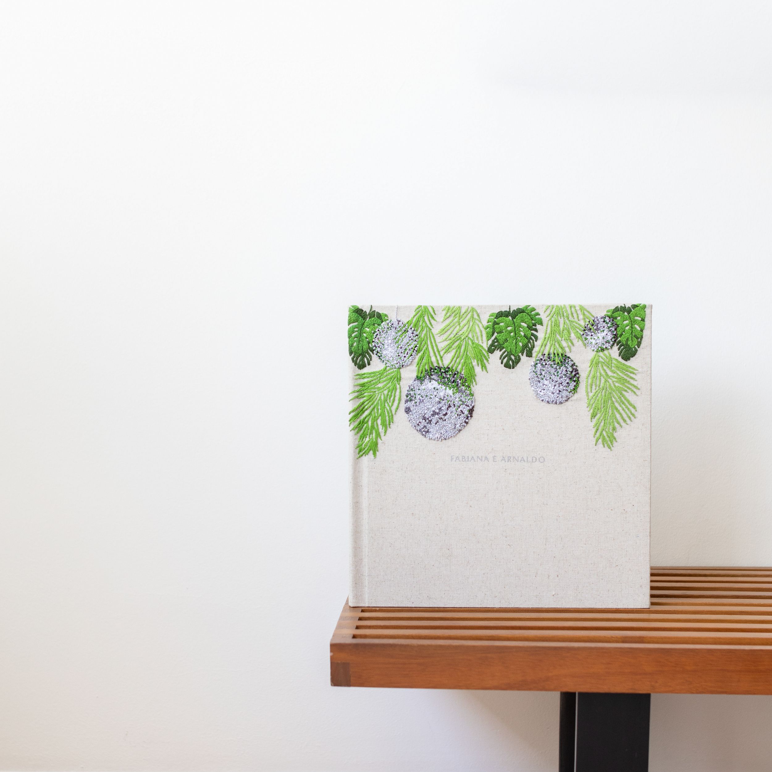 A minimalist square photo album cover with pine needles and ornaments design along the top edge on a wooden shelf.
