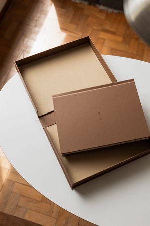 Open brown cardboard box with lid on white table, parquet wood floor background, natural light.