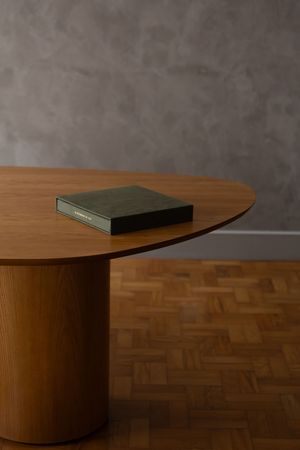 A round wooden dining table with a cylindrical pedestal base, topped with a green book, on a parquet floor.