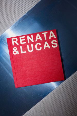 Red hardcover book titled Renata & Lucas placed on a blue metallic surface with diagonal lines.