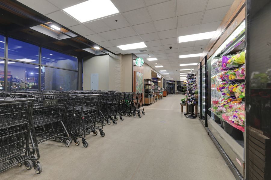 Interior of a grocery store showing rows of shopping carts near large windows and refrigerated display cases along the aisle.