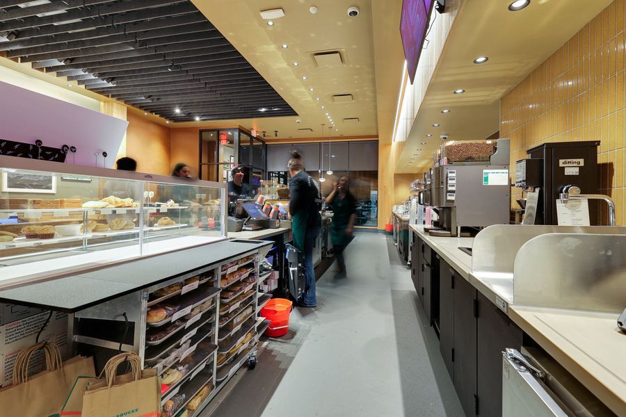 Busy cafe counter with display cases, baked goods, and customers ordering at a modern food service area with warm lighting.