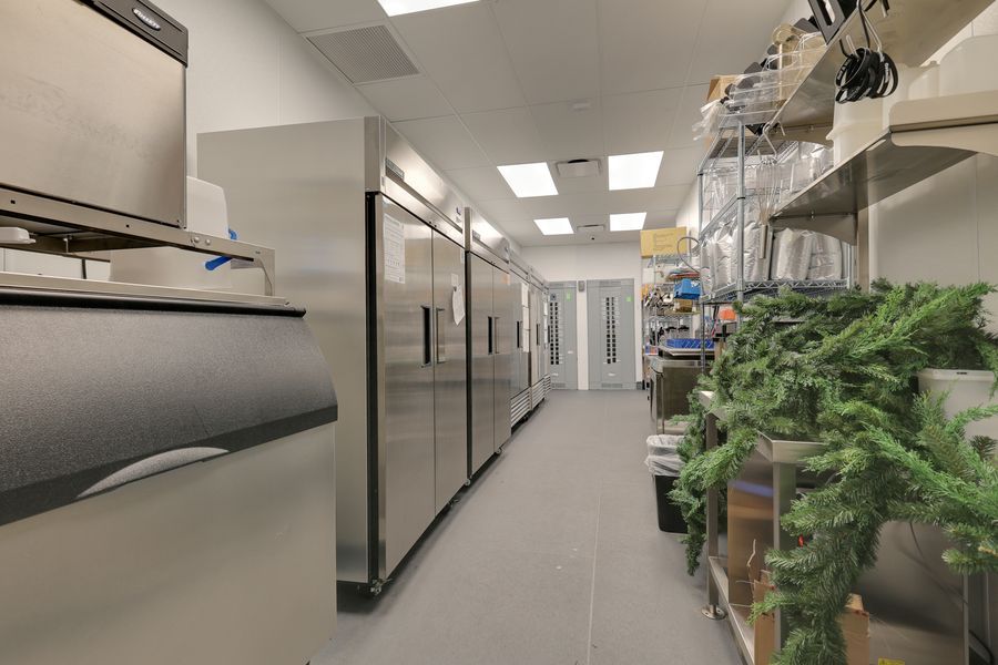 Stainless steel commercial kitchen with large refrigerators and storage shelves lined with artificial greenery.