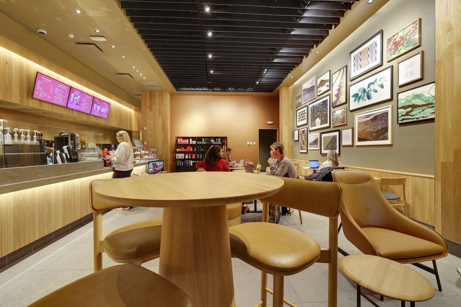 Modern coffee shop interior with wooden furniture, round tables, and framed artwork on warm terracotta walls.