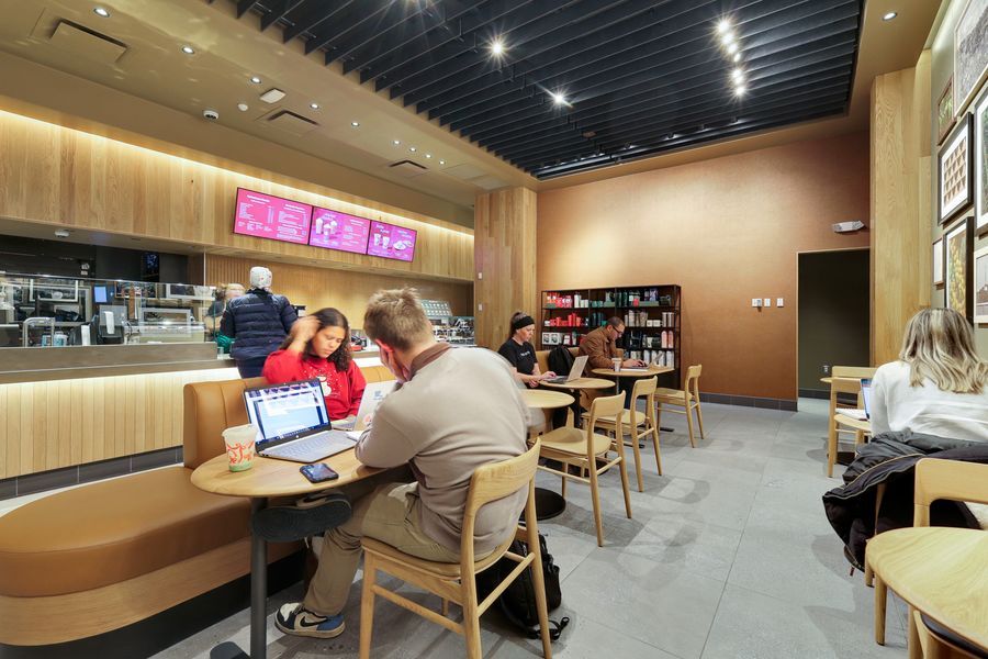 Modern cafe interior with students working on laptops at wooden tables near a service counter with digital menu boards.