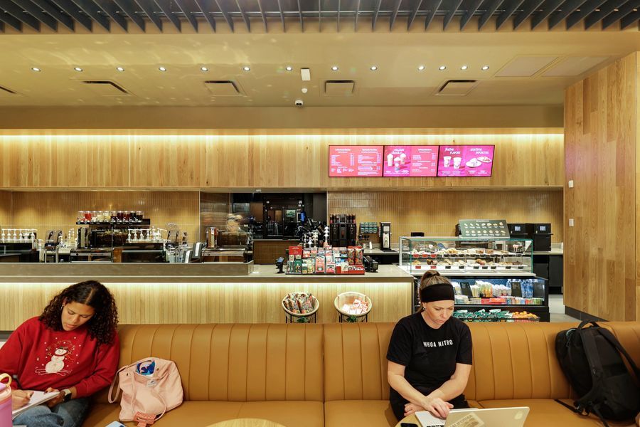 Starbucks cafe interior with customers seated at booth, illuminated menu boards, and barista counter with pastry display.