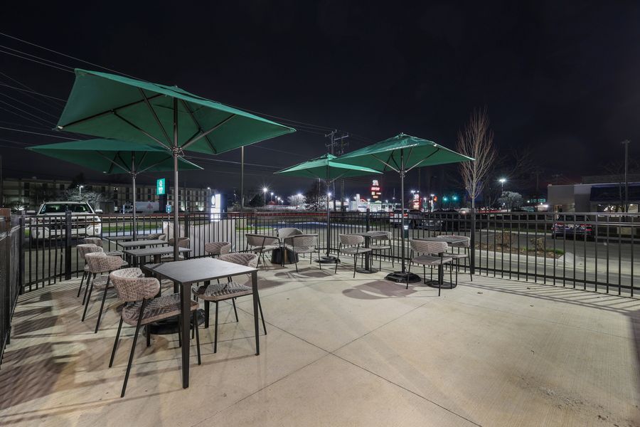 Outdoor patio with green umbrellas and metal chairs at night, overlooking a parking lot with city lights in the background.